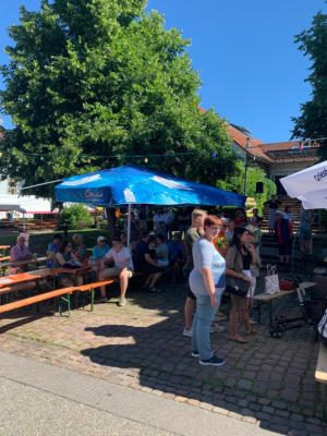 Gäste sitzen an Biertischen unter einem blauen Sonnenschirm, im Vordergrund stehen Frauen in der Sonne; dahinter ein großer Baum und blauer Himmel.