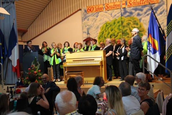 Chor mit hellgrünen Schals auf der Bühne hinter einem hellen Klavier, darüber das Banner „60 Jahre Soudron – Schluttenbach“, seitlich Fahnen; links ein Redner am Pult, vorne Publikum an Tischen.
