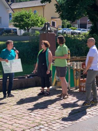 Ein Mann im türkisfarbenen Poloshirt spricht ins Mikrofon und hält einen großen Spendenscheck. Neben ihm zwei Frauen in Grün und ein Mann im weißen Hemd, dahinter Brunnensäule und Ausschank.