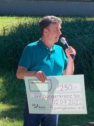 Ein Mann im türkisfarbenen Poloshirt spricht ins Mikrofon und hält einen Spendenscheck über 250 Euro für den GV Sängerkranz, ausgestellt vom Pfennigbasar e.V. am 02.07.2022.