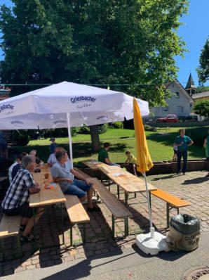 Gäste an Biertischen unter einem großen weißen Sonnenschirm, daneben ein zugeklappter gelber Schirm; im Hintergrund Wiese, Bäume, Häuser und ein Turm.