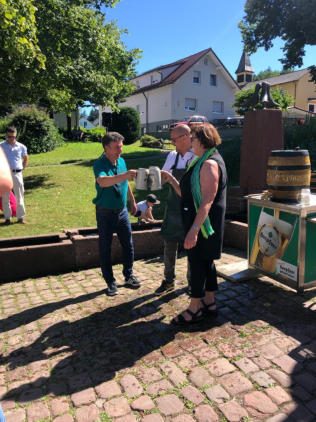 Zwei Männer stoßen mit Steinkrügen an, einer im türkisfarbenen Poloshirt, einer mit dunkler Schürze; daneben eine Frau mit hellgrünem Schal. Hinten Brunnentrog, Bierfass, Wiese und Häuser.